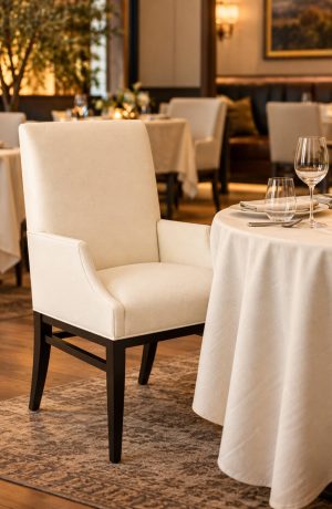 Cream upholstered dining armchair with a high back, curved arms, and dark wood legs beside a table in an upscale restaurant